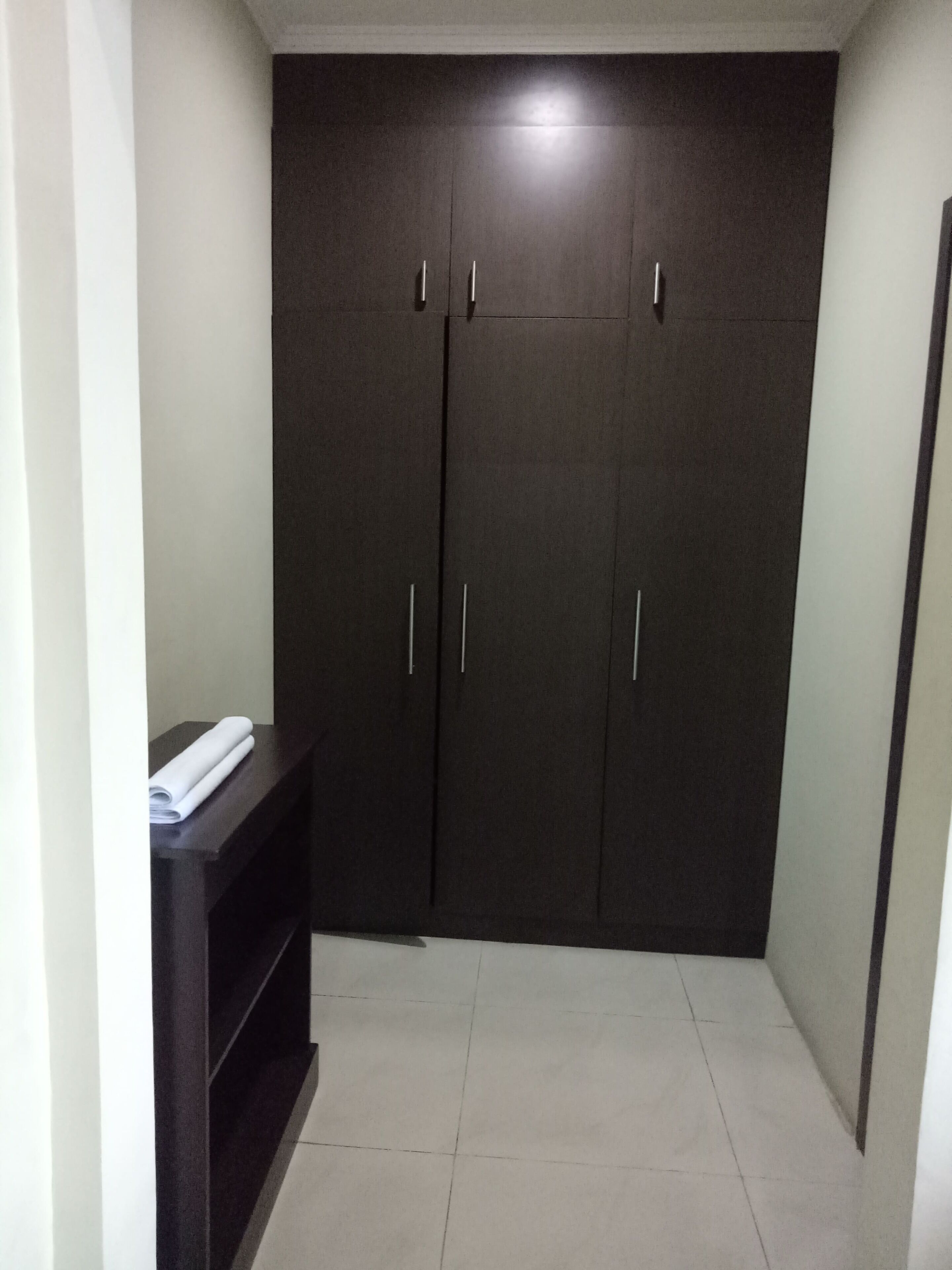 A room with a dark-colored wardrobe, a small shelf with a rolled-up white item, and a tiled floor.