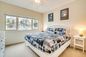 Une chambre avec un grand lit, une fenêtre avec des stores, un ventilateur de plafond et deux tableaux encadrés accrochés au mur.