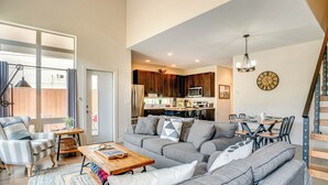 Ein geräumiges Wohnzimmer mit grauem Sofa, einem Holztisch und einem gestreiften Sessel. Im Hintergrund ist eine Küche mit dunklen Holzschränken und einer Mikrowelle zu sehen.