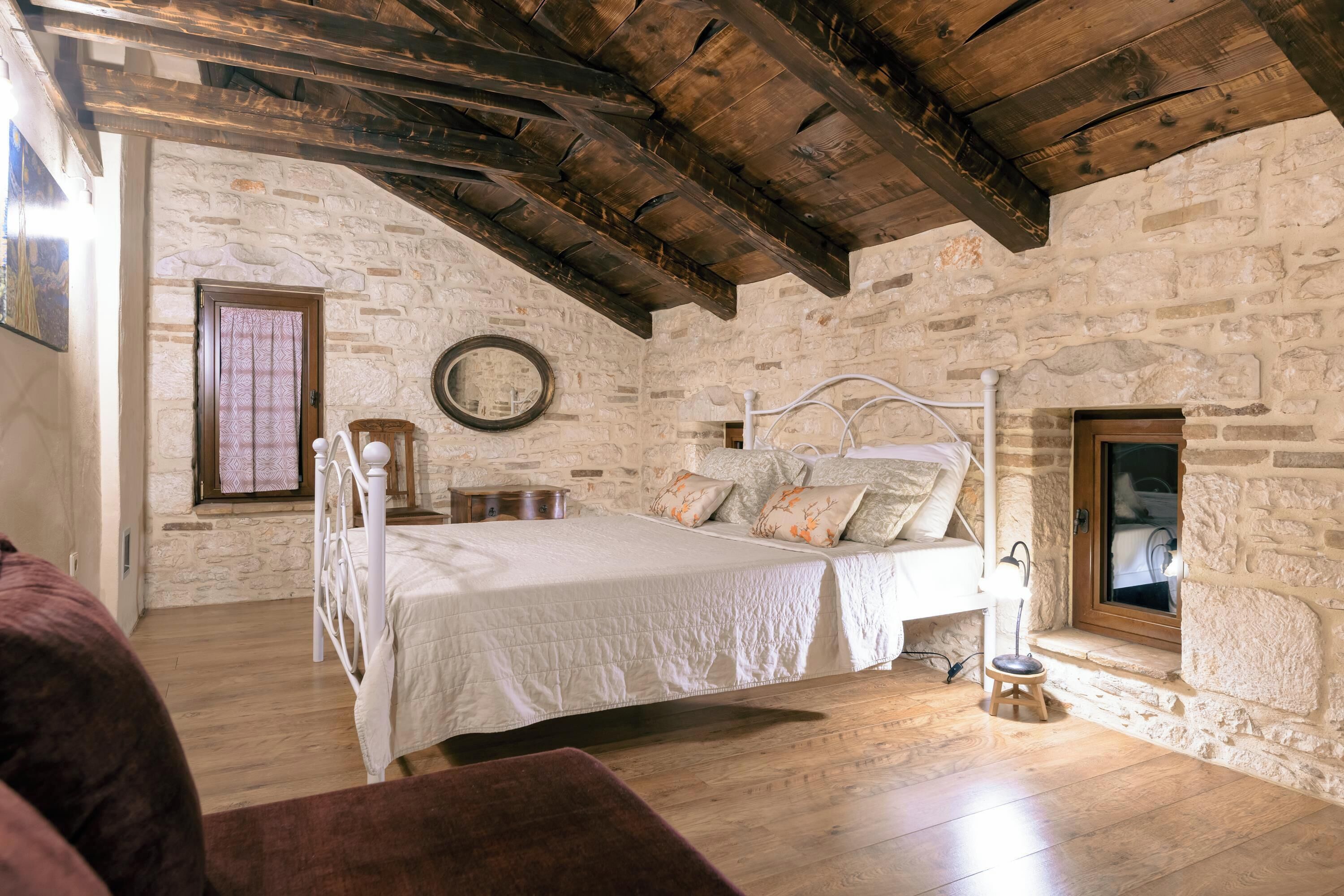 Une chambre avec un mur en pierre, des poutres en bois, un lit, une table de chevet, un miroir et une fenêtre.