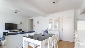Un salon moderne comprenant un canapé, une table à manger avec un plateau en verre, des chaises blanches, un téléviseur et un meuble avec des portes en verre.