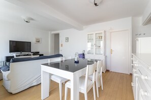 Un salon moderne comprenant un canapé, une table à manger avec un plateau en verre, des chaises blanches, un téléviseur et un meuble avec des portes en verre.
