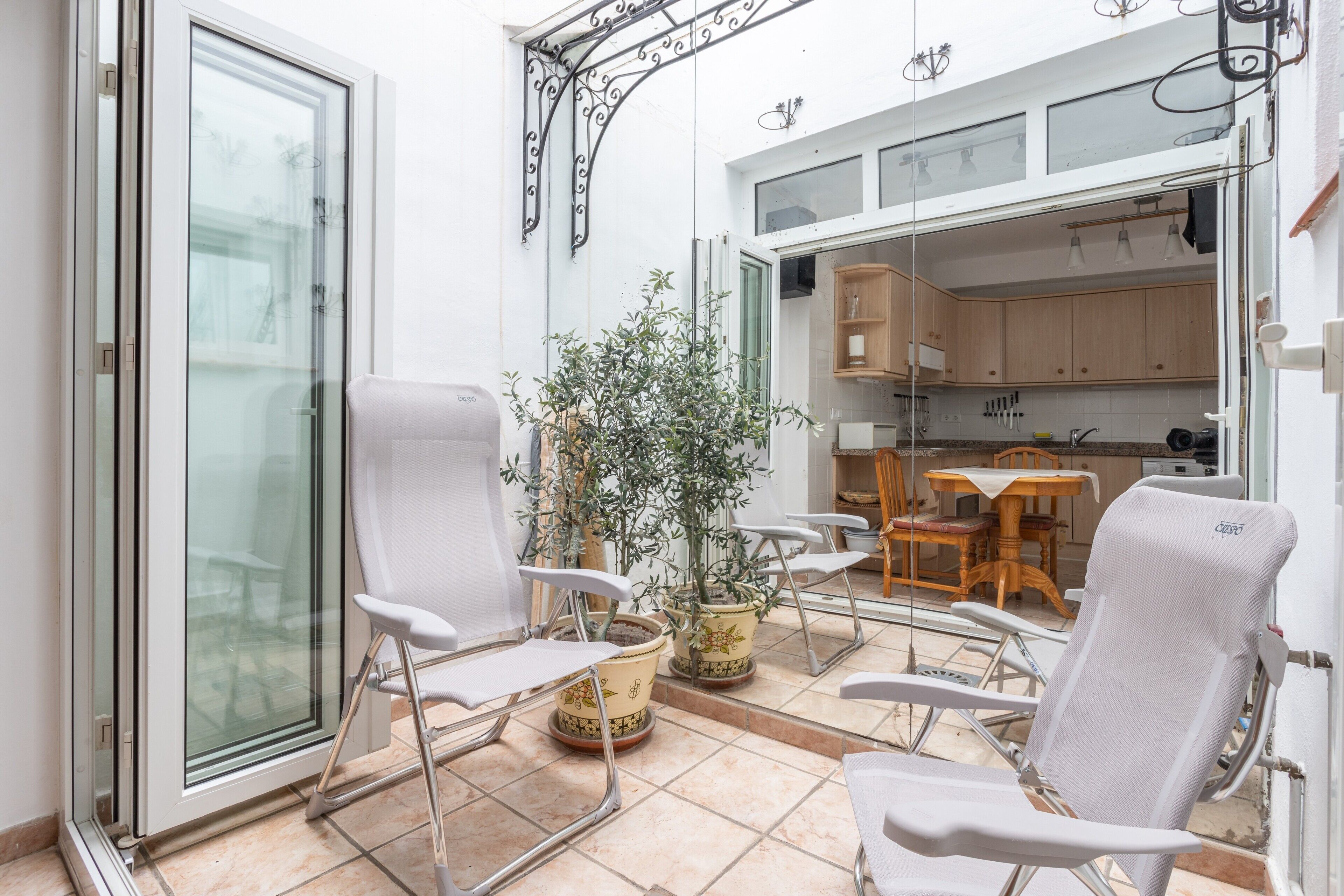 A sunlit patio with white chairs, a potted plant, and a view of a kitchen through an open door.
