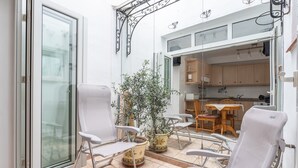 Eine sonnendurchflutete Terrasse mit weißen Stühlen, einem Topfpflanzen und Blick auf eine Küche durch eine offene Tür.