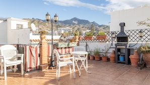 Eine Dachterrasse mit Gartenmöbeln, Blumentöpfen, einem Grill und Blick auf Berge.