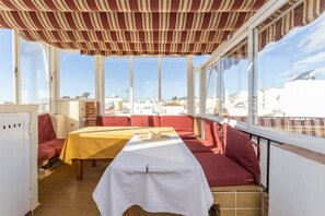 Une terrasse ensoleillée avec une table et des sièges rouges, de grandes fenêtres et une vue sur les bâtiments.