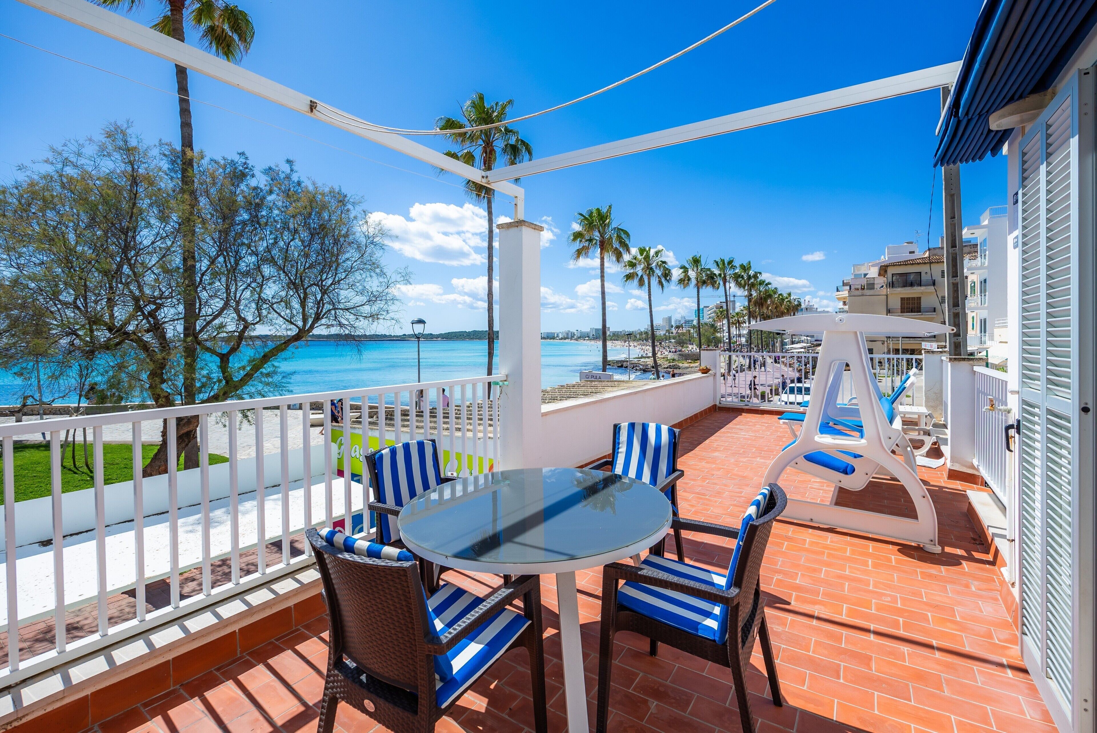 A balcony with a glass table and chairs, overlooking a beach and palm trees.