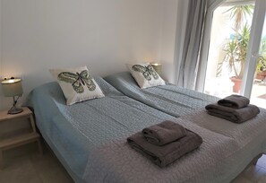 Une chambre avec deux lits, une table de chevet avec une lampe, et une vue sur les palmiers à l’extérieur.