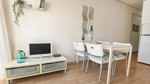 Un'area pranzo moderna con tavolo e sedie bianche, una TV su un mobile in legno e una pianta su uno scaffale.