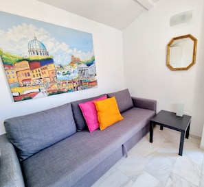 A modern living room with a grey sofa, colorful cushions, a painting of a cityscape, a gold-framed mirror, and a small black table.