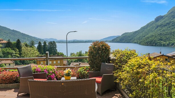 Una terraza con mobiliario de mimbre, una mesa con una maceta amarilla y vistas a un lago rodeado de colinas.