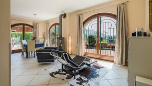 Una sala de estar con sofá, una mesa de comedor y televisor. Amplias ventanas con cortinas, y vistas a un jardín.