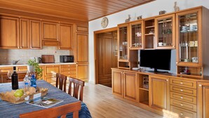 Keittiössä on puukehikoiset kaapit, ruokapöytä tuoleineen, televisio ja seinällä oleva kello.
