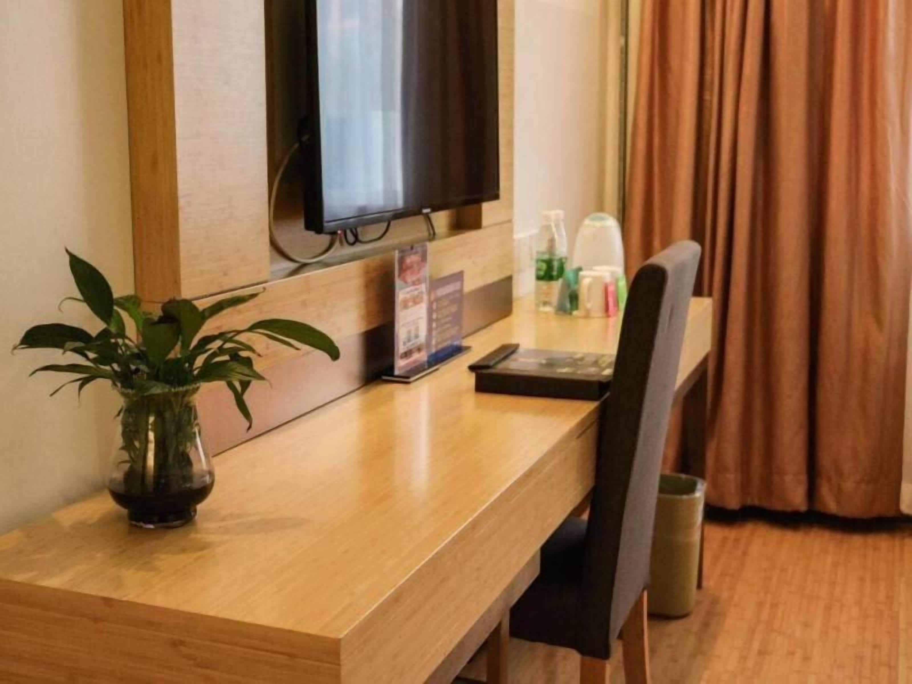 Une chambre d’hôtel avec un bureau en bois, un téléviseur fixé au mur, une plante en pot et une fenêtre avec des rideaux.