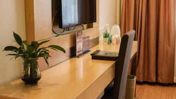 Une chambre d’hôtel avec un bureau en bois, un téléviseur fixé au mur, une plante en pot et une fenêtre avec des rideaux.