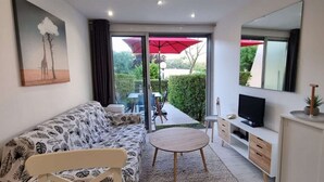 A modern living room with a sofa, a small round table, a TV, and a cabinet. There is a glass door leading to an outdoor area with a red umbrella. - Pretty T2 garden level at Couchant 200m from the beach in a residence with swimming pool (La Grande-Motte)