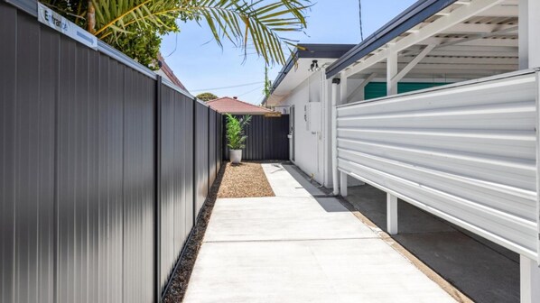 A narrow pathway leading to a garage with a white roller door, flanked by dark grey fences and a small garden area with a palm tree. - Bellara Springs - walk to the water (BELLARA)
