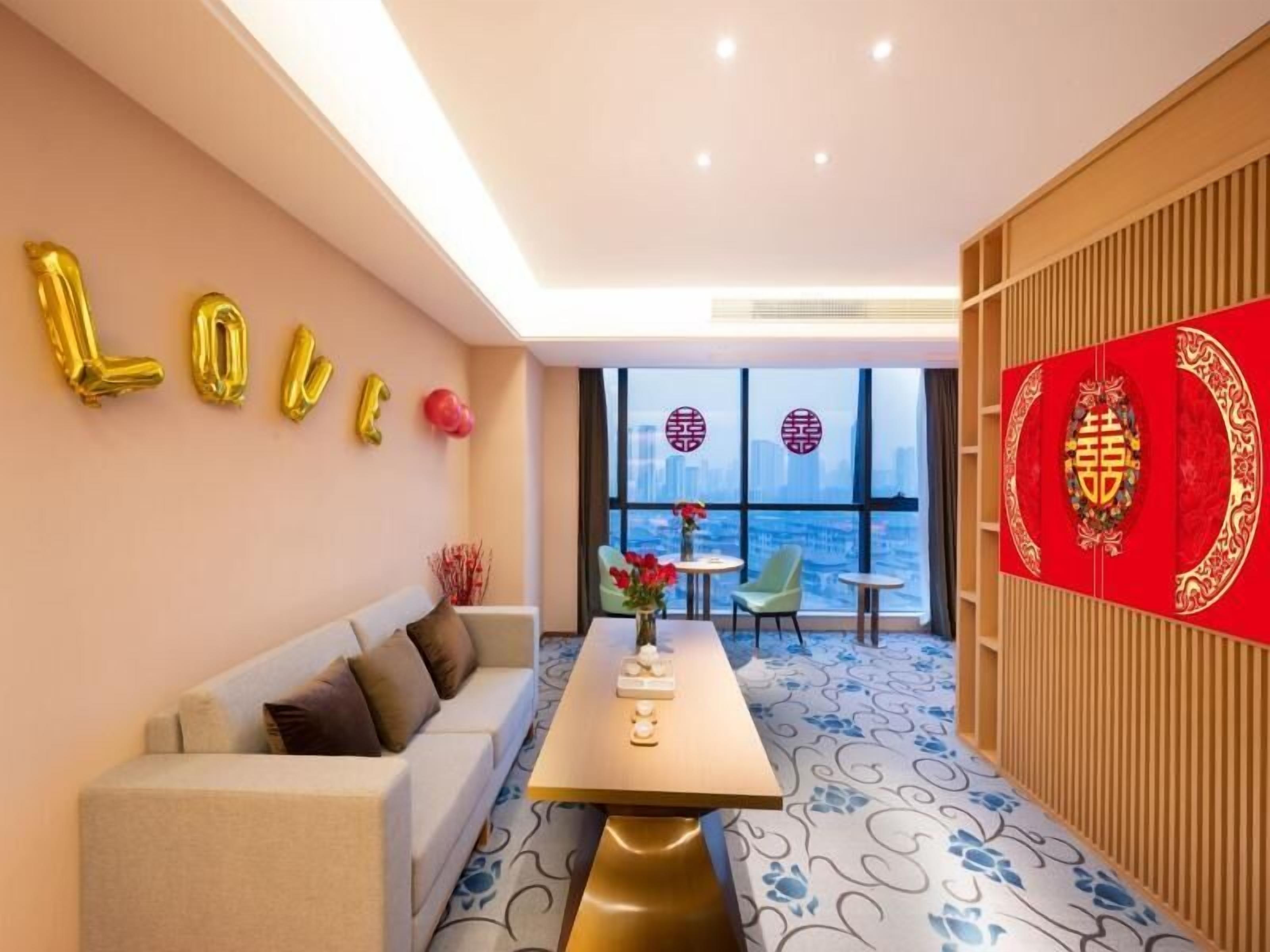 A modern hotel room with a sofa, a dining table, and a view of the city through large windows.