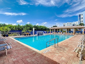 A swimming pool with lounge chairs and a covered area, surrounded by a blue fence and greenery.