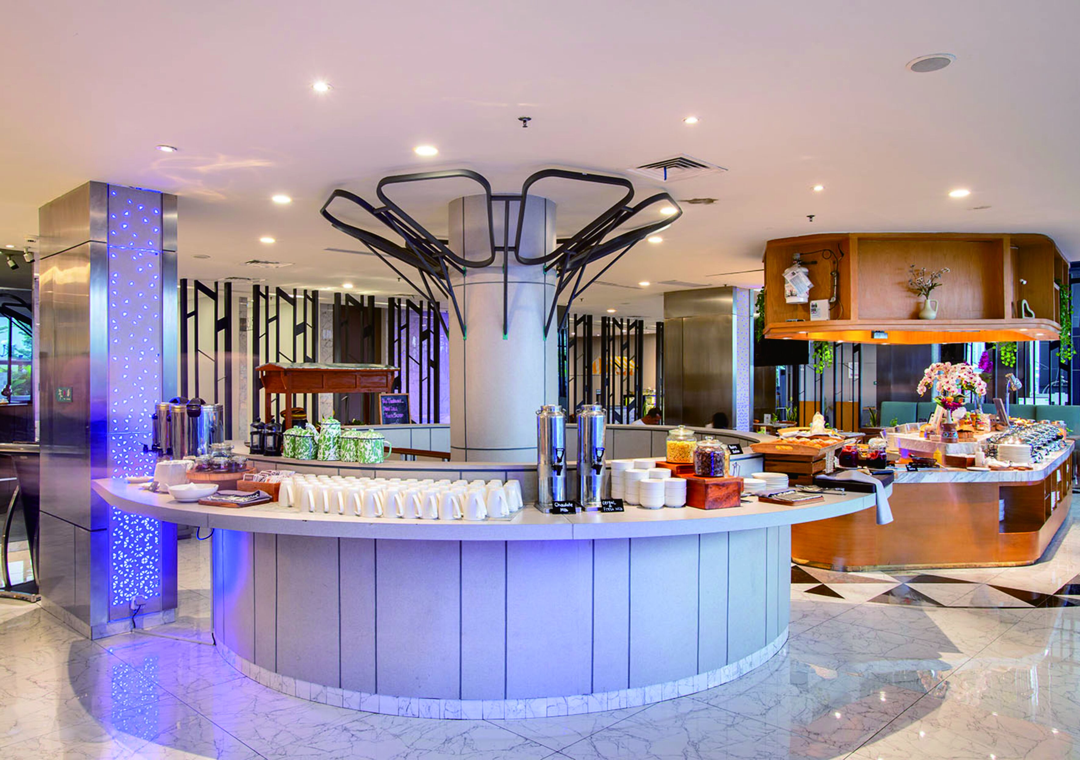 Un moderno vestíbulo de hotel con una zona de bar circular central que ofrece una variedad de dispensadores de comida y bebidas, arreglos de asientos y una estructura decorativa en el techo.
