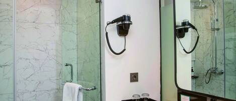 A modern bathroom with a glass shower enclosure, a white marble wall, a black hairdryer mounted on the wall, and a large mirror above the sink.