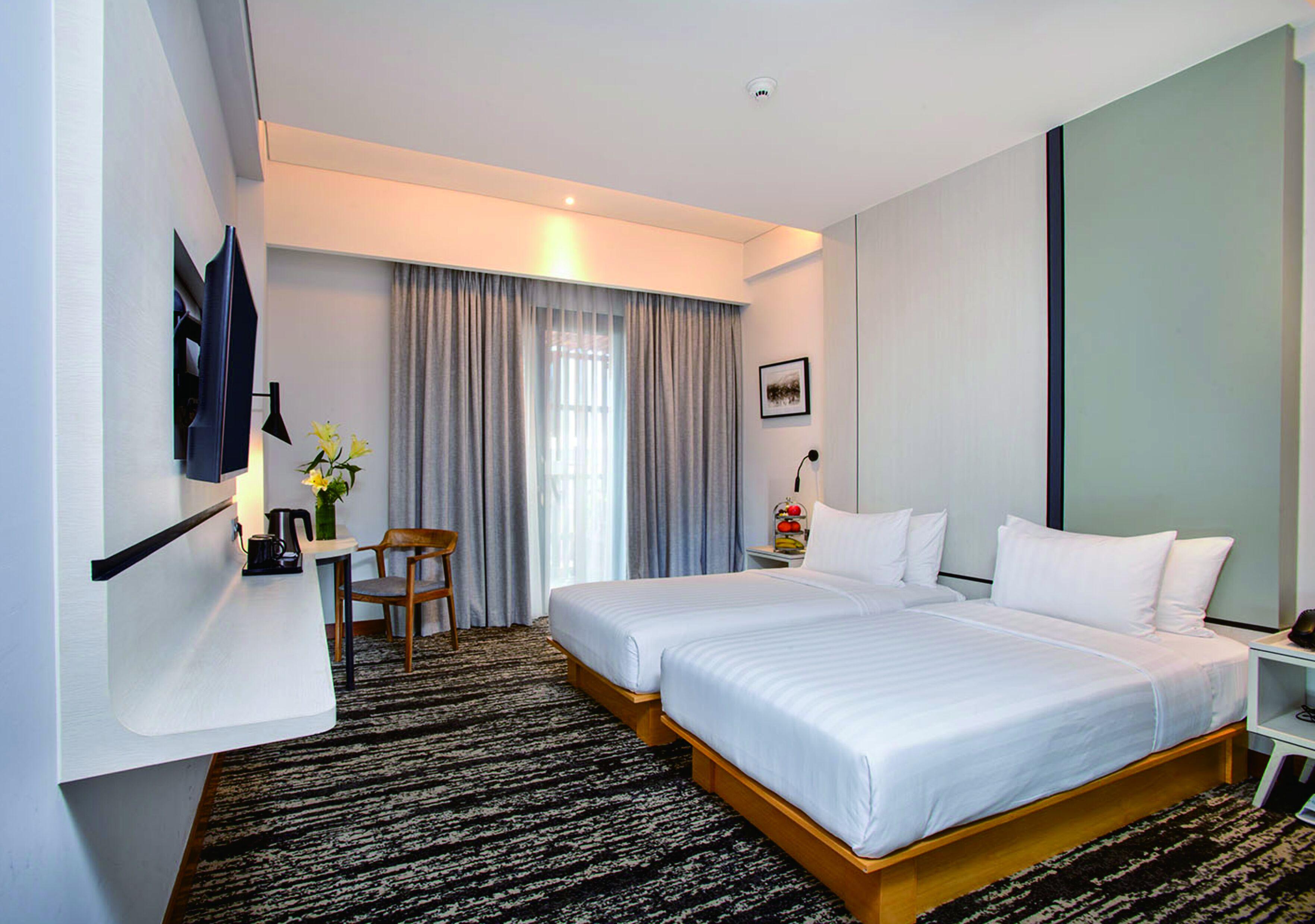 A hotel room with a double bed, a desk with a chair, a TV mounted on the wall, and a window with curtains.