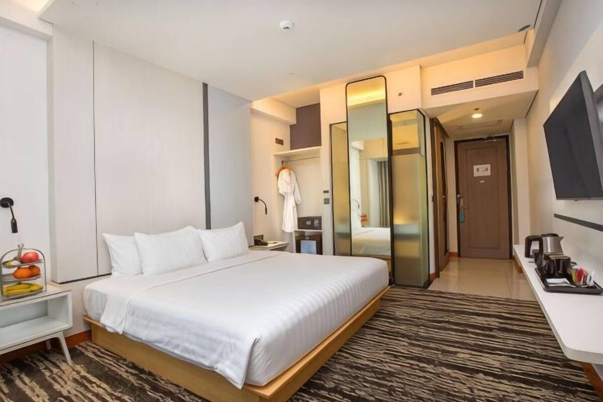 A modern hotel room with a large bed, a bedside table with a fruit bowl, a mirror, a TV, and a bathroom with a shower and toilet.