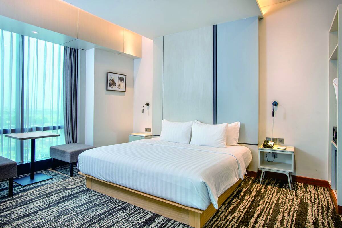 A hotel room with a large bed, a desk, a chair, and a view of the outdoors through the window.
