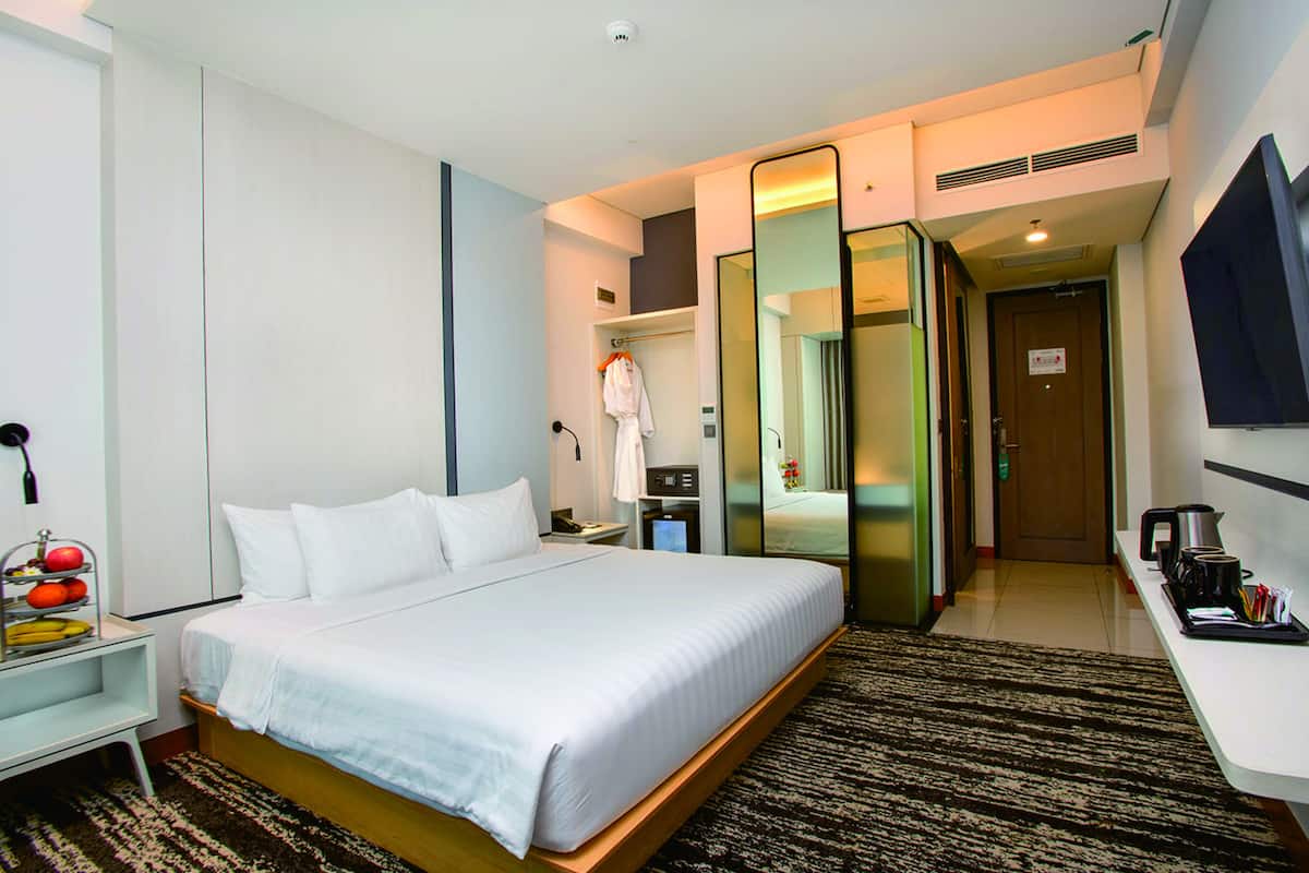 A hotel room with a large bed, a bedside table with a lamp, a mirror, a wardrobe, a TV, and a coffee maker.