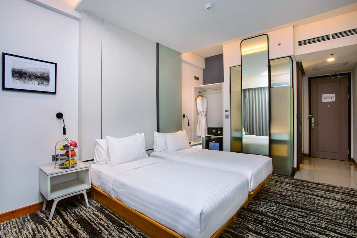 A hotel room with two beds, a bedside table with a fruit basket, a mirror, and a robe hanging on a rack.