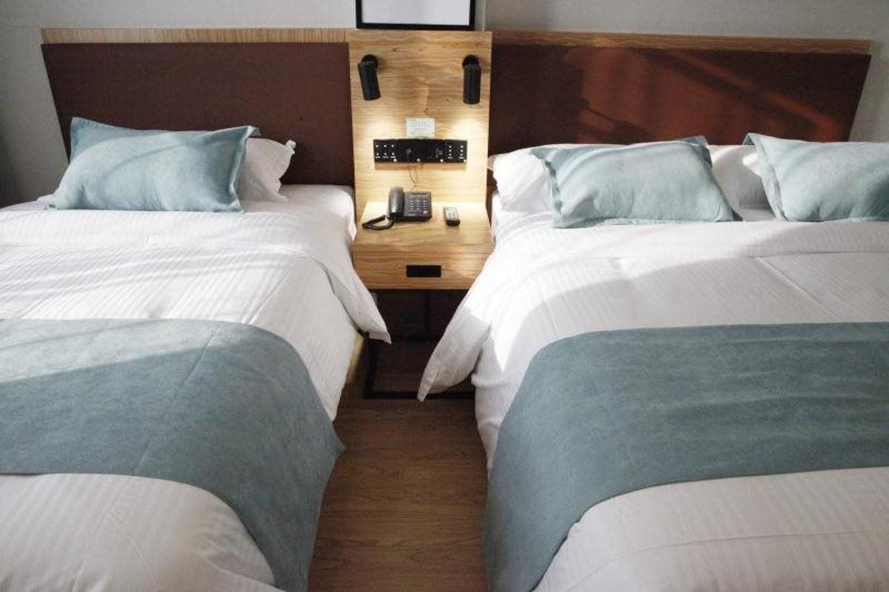 Habitación de hotel con dos camas, cabecero de madera, mesita de noche con teléfono y lámpara de pared.