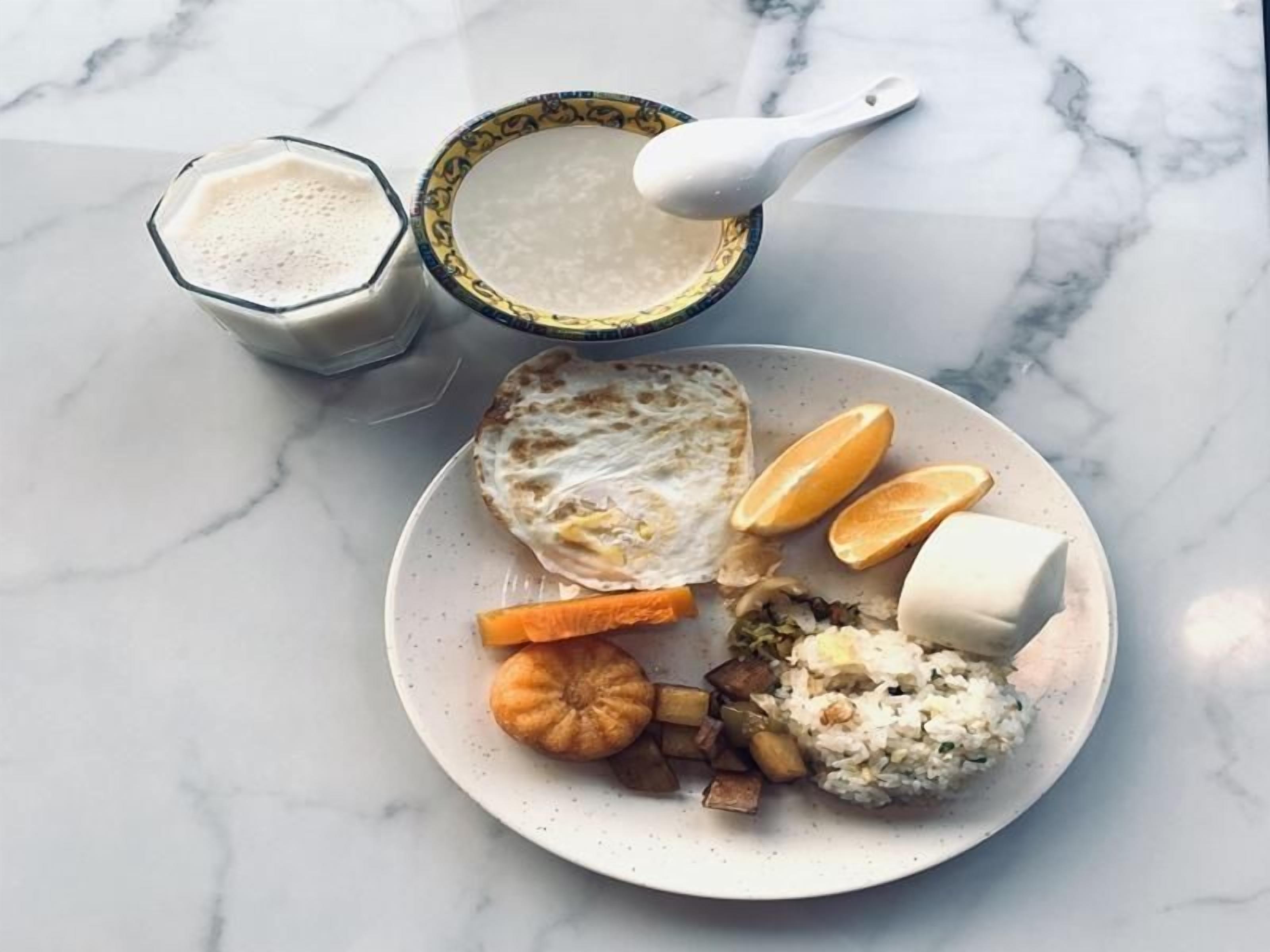 A plate with toast, orange slices, a dollop of butter, a small bowl of soup, and a glass of milk on a marble surface.