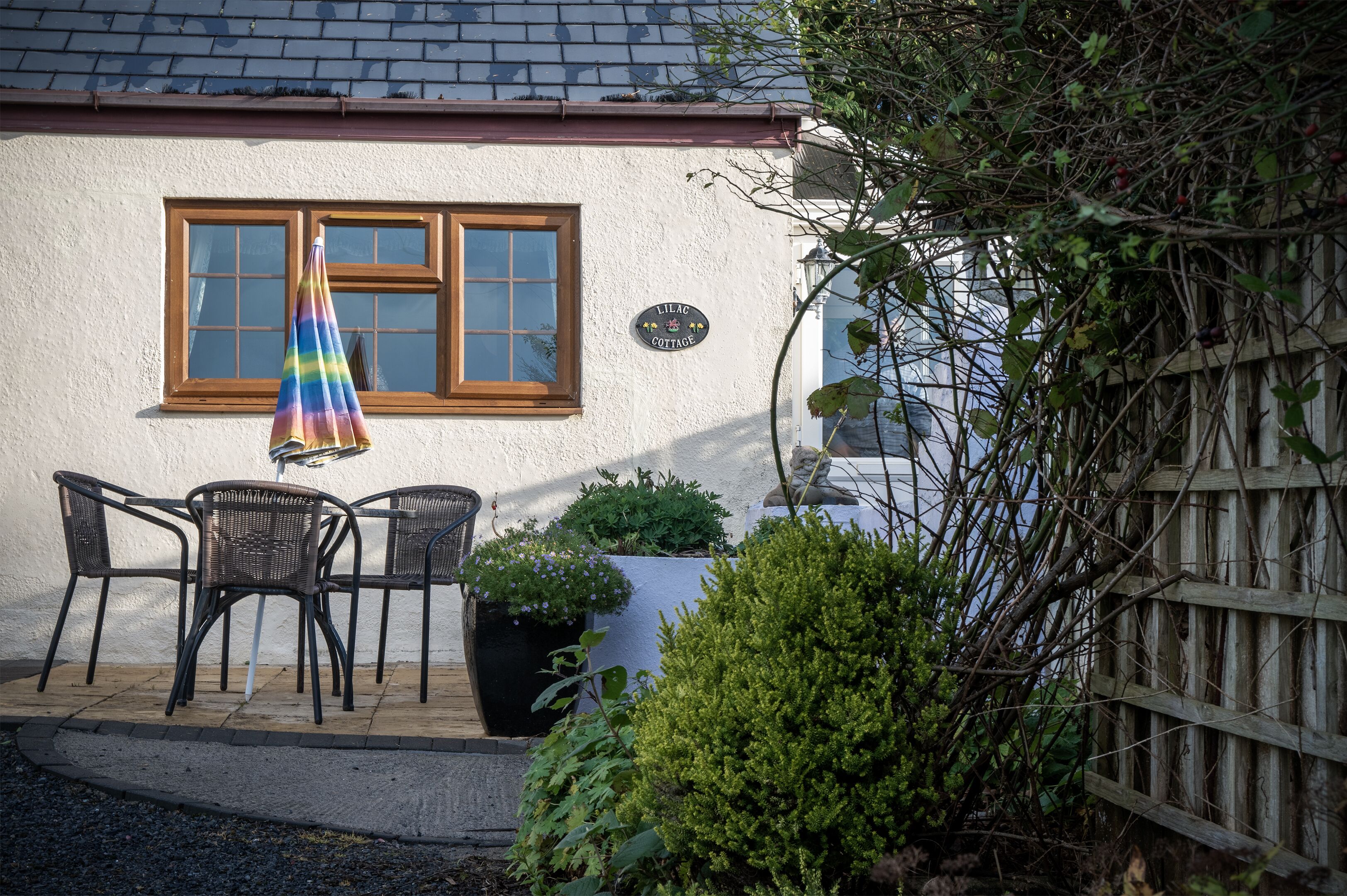 A cottage with a wooden window, a sign reading "LILAC COTTAGE," and a patio with chairs and a potted plant.