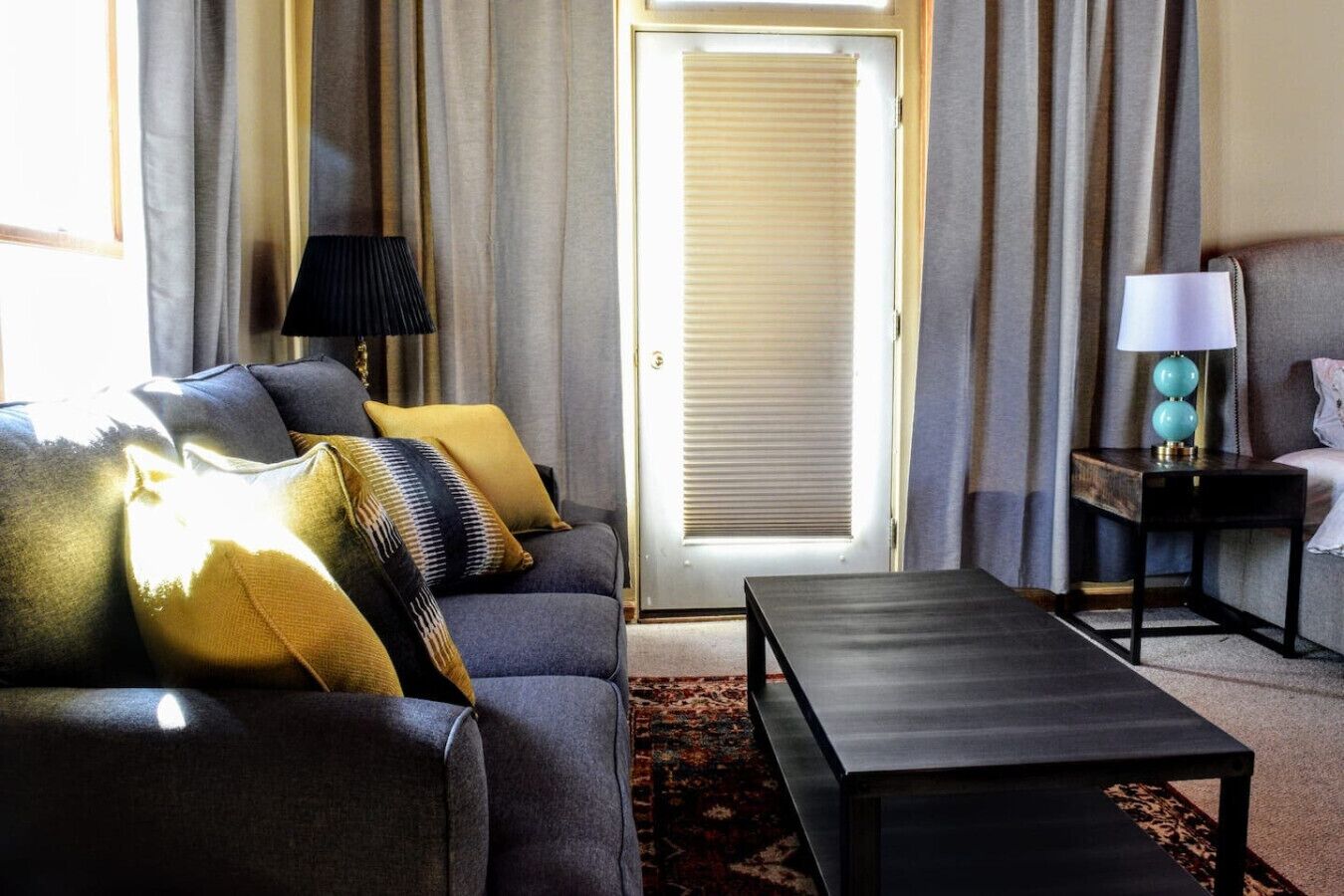 A modern living room with a grey sofa, patterned cushions, a black coffee table, a side table with a lamp, and a view through a door with curtains.