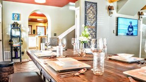Un comedor con una larga mesa de madera dispuesta para una comida, una cocina al fondo y un adorno en la pared.
