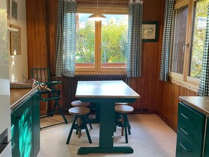 A kitchen with a wooden table and stools, checkered curtains, and a view of greenery outside. - Spacious chalet in Saint-Gervais, 7 ppl, Mont-Blanc view (Saint-Gervais-les-Bains)