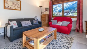 A living room with a dark grey sofa, a red armchair, a wooden coffee table, and a view of snow-capped mountains through a window with plaid curtains.