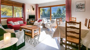 A spacious living room with a red sofa, a wooden coffee table, and a dining table with chairs. There are large windows offering a view of trees and mountains.