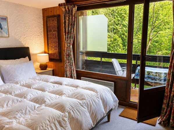 A bedroom with a large bed, a nightstand with a lamp, a window with curtains, and a view of greenery outside.
