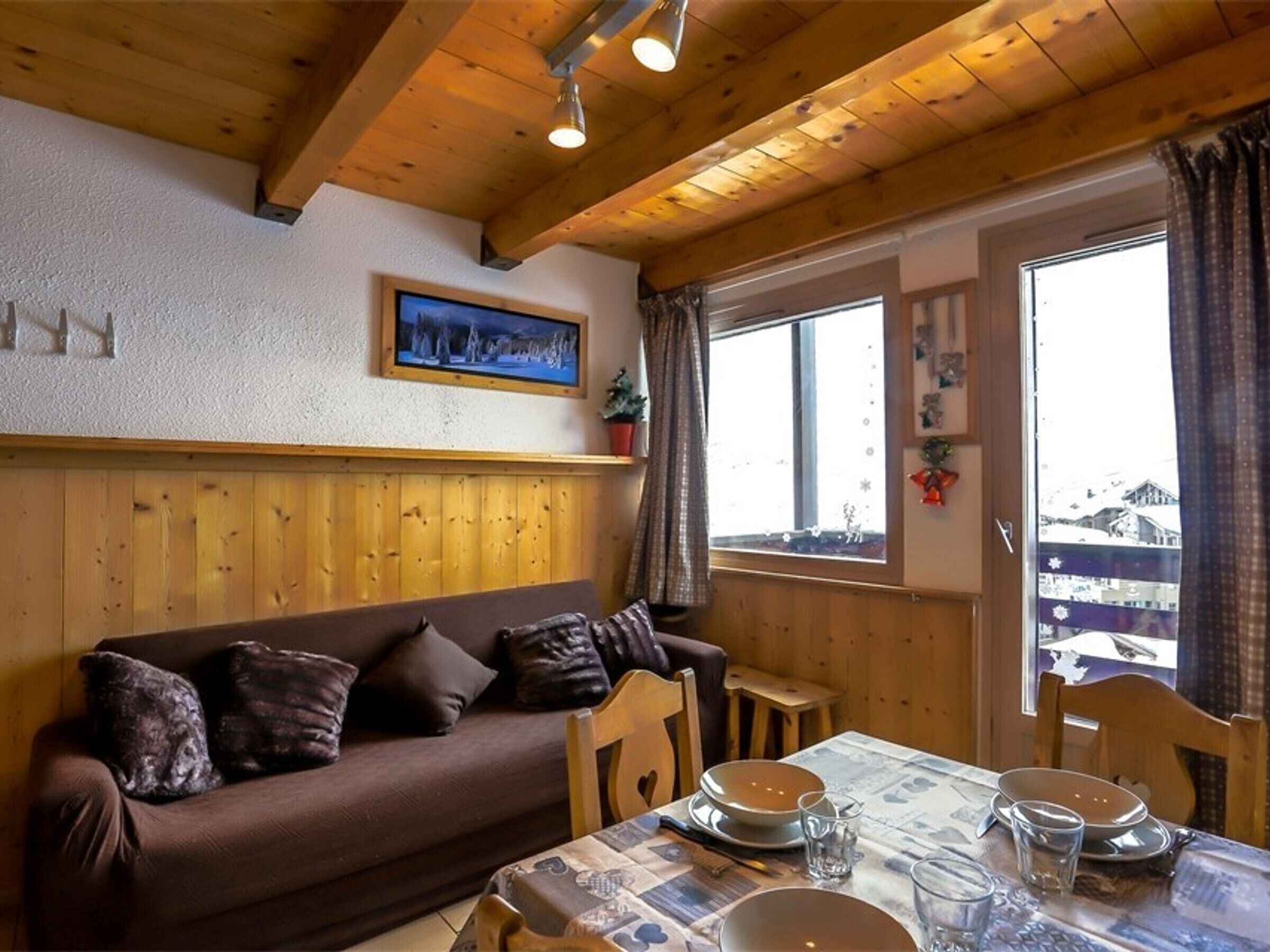 Una habitación acogedora con techo de madera, un sofá marrón con cojines, una mesa de comedor con platos y vasos, y una ventana con cortinas.
