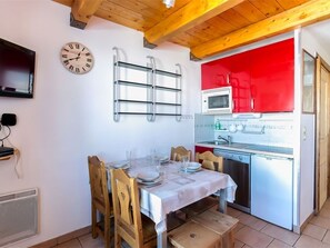 Uma sala de jantar com uma mesa de madeira posta para quatro pessoas, um relógio na parede e uma cozinha com armários vermelhos.