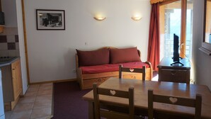 Une pièce chaleureuse avec une table et des chaises en bois, un canapé rouge, un petit coin cuisine et une fenêtre avec des rideaux.