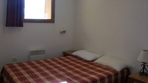 Une chambre avec un lit, une fenêtre avec des rideaux, une table de chevet avec une lampe et un radiateur fixé au mur.