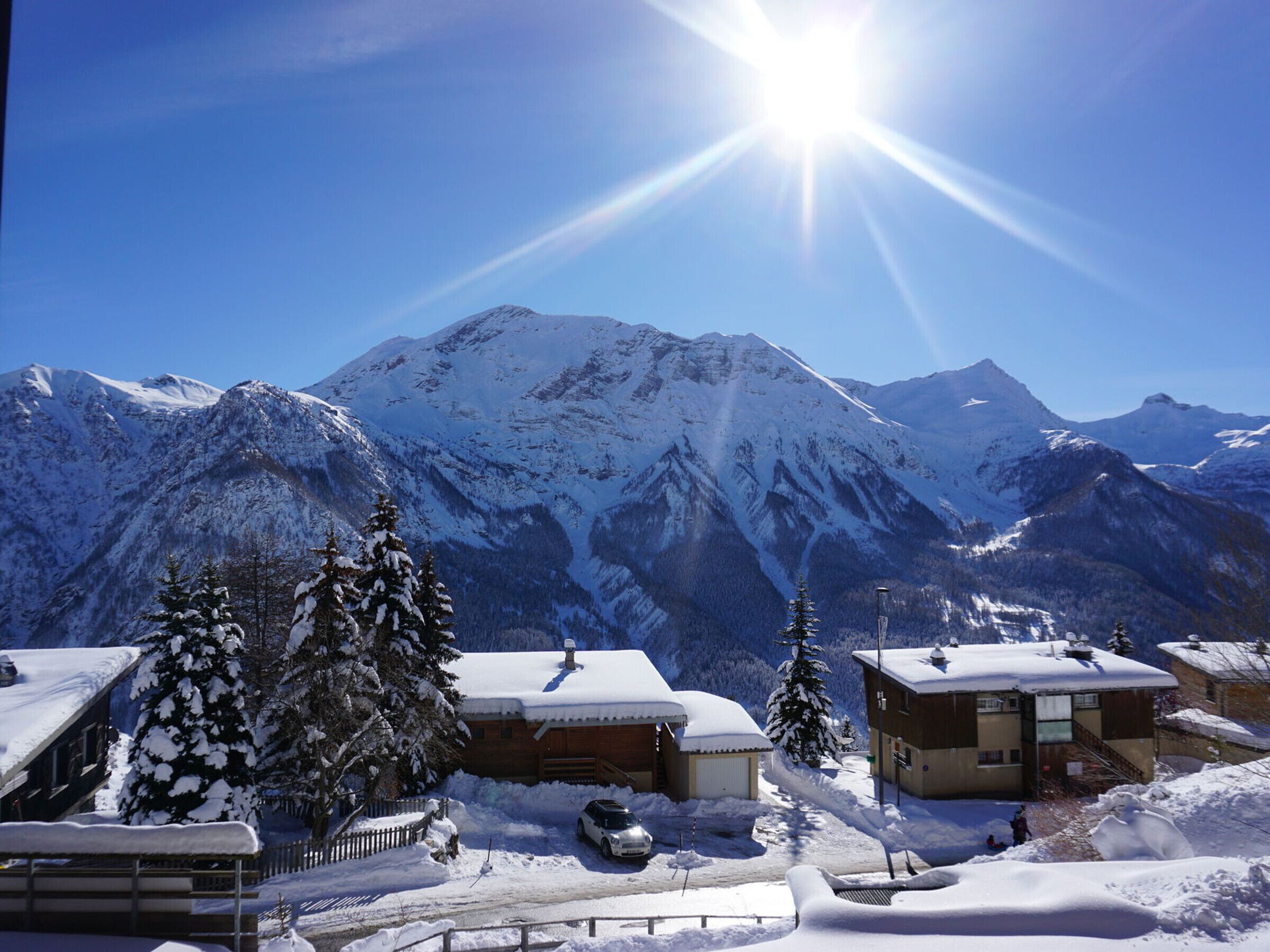 Un pueblo montañoso cubierto de nieve, con edificios y árboles, un cielo despejado y un sol brillante.