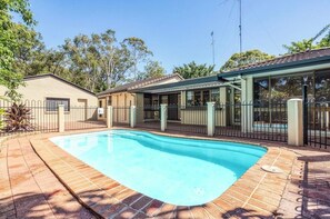 A swimming pool with a tiled deck, surrounded by a fence and a covered area with glass walls.