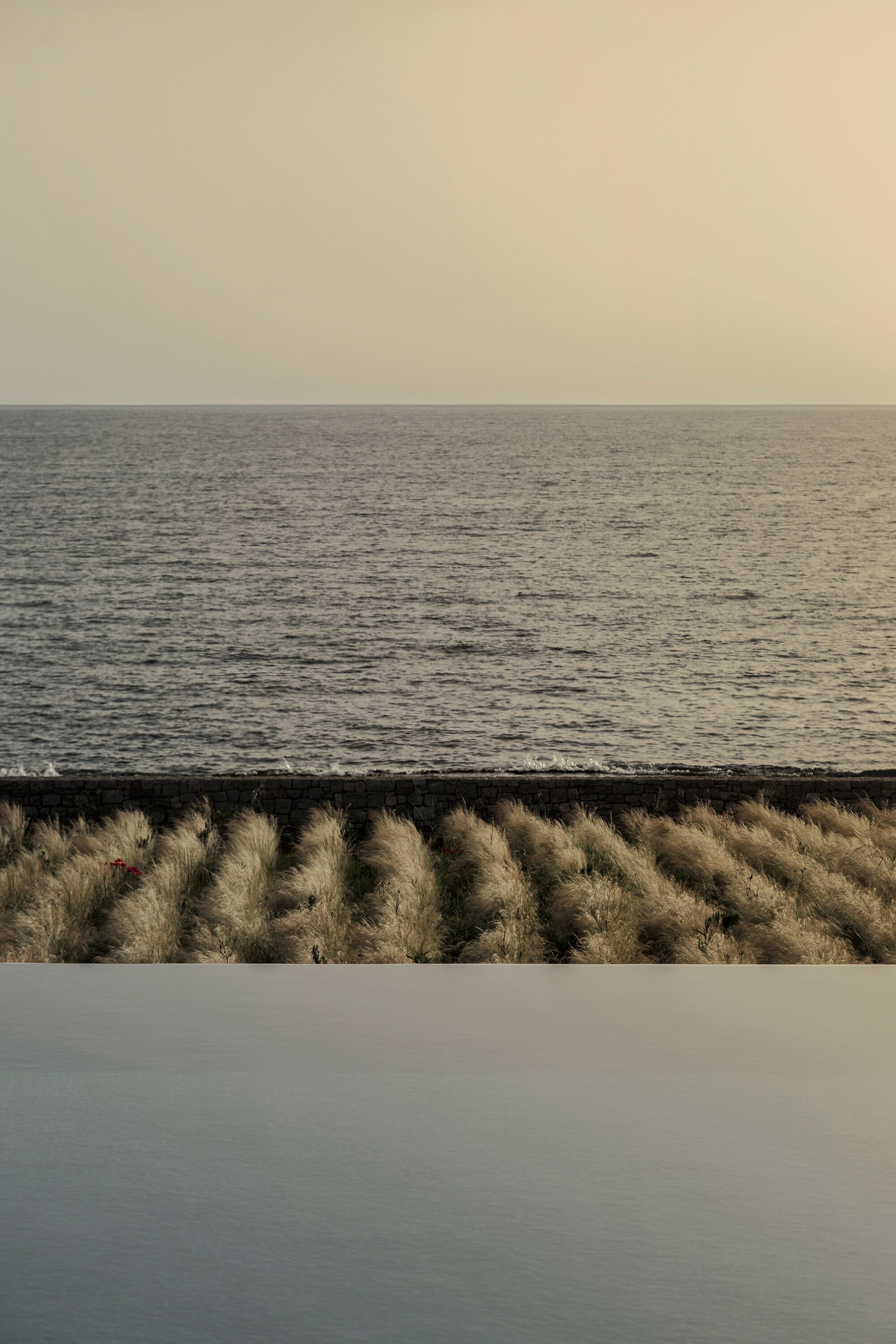 A calm sea with a stone wall and grass in the foreground.