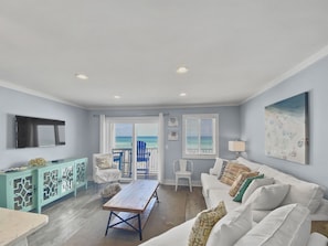 Uma sala de estar ampla com uma grande janela que oferece vista para o oceano, um sofá branco, uma mesa de centro de madeira e um console azul-turquesa com objetos decorativos.