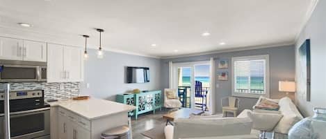 A modern kitchen with white cabinets, a central island, and stainless steel appliances. There is a dining area with a white table and chairs, and a living room with a white sofa and a glass coffee table.