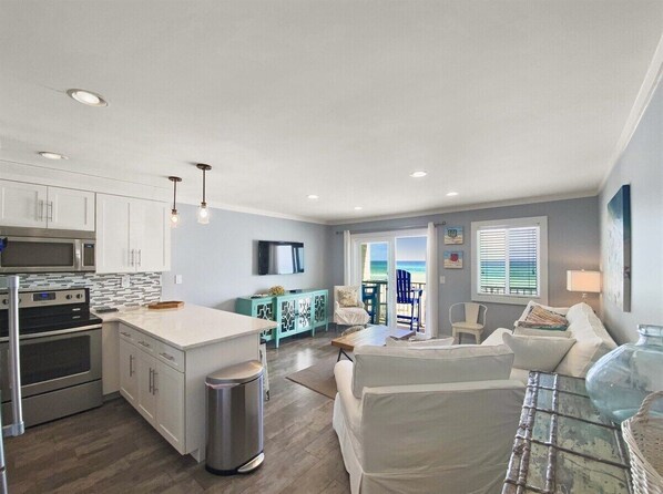A modern kitchen with white cabinets, a central island, and stainless steel appliances. There is a dining area with a white table and chairs, and a living room with a white sofa and a glass coffee table.