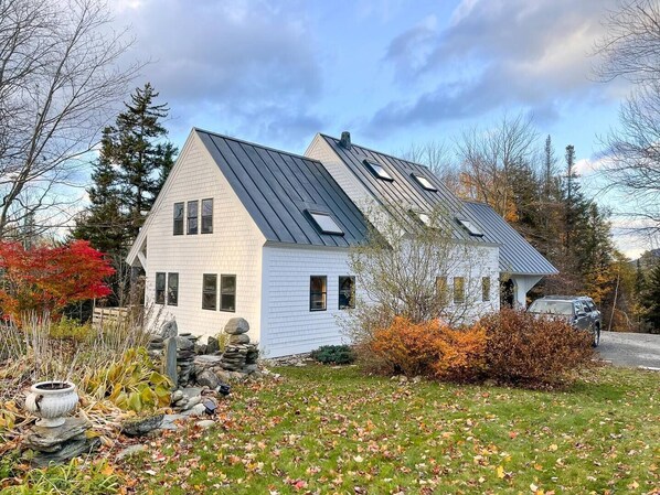 A white house with a sloped roof and multiple windows, surrounded by autumn foliage and a stone wall. - Hand Crafted Post and Beam Home with Mountain Views Minutes from Sugarbush (Warren)